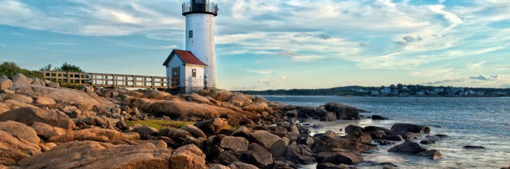 A white lighthouse with a red-roofed building stands on rocky terrain by the sea, looking much like a serene mental health retreat. A wooden walkway leads you there, while the partly cloudy sky gently embraces distant houses lining the opposite Boston shore.