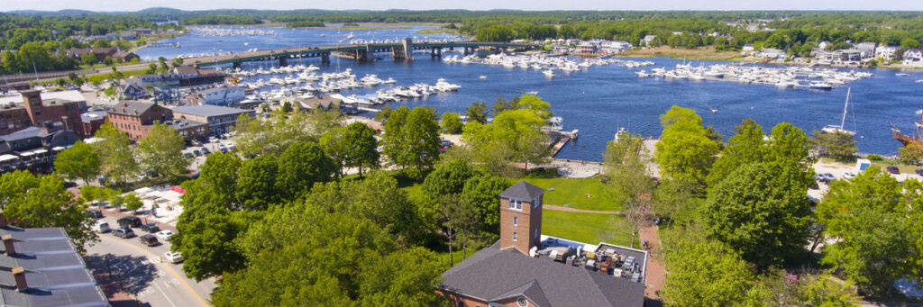 Aerial view of a coastal town in Plymouth County, featuring a river and marina filled with boats. Amidst green trees, buildings, and a bridge under the clear blue sky, the serene setting reflects tranquility often found near mental health rehabs.