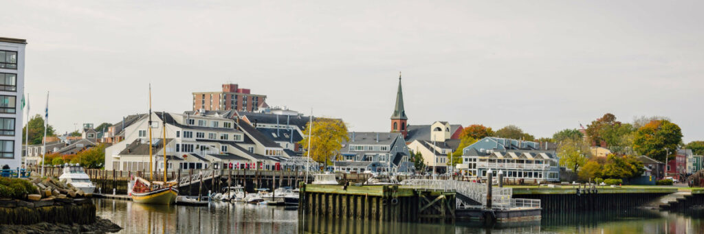 A scenic coastal town, reminiscent of a mental health retreat in Boston, features a harbor with docked sailboats, quaint buildings, and a church with a tall steeple. Trees with autumn foliage add color as the calm water reflects the picturesque setting on a clear day.