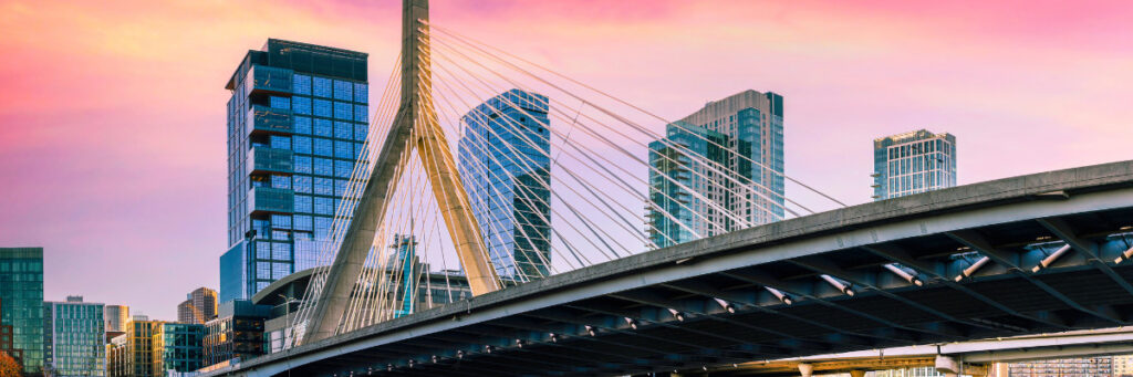 A modern cable-stayed bridge with a sleek design stretches gracefully against an urban skyline adorned with contemporary skyscrapers. The sky, vibrant with pink and orange hues, mirrors the hope found in mental health treatment in Worcester County at both sunrise and sunset.