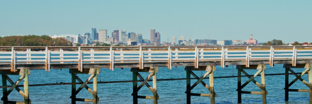 A wooden pier stretches over calm blue water with a distant city skyline in the background, evoking a serene escape akin to the tranquility found at mental health rehabs. The sky is clear and blue, with trees flanking the scene on the left, reminiscent of peaceful retreats near Waltham.
