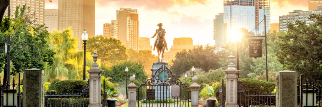 A statue of a person on horseback stands at the entrance of the park, surrounded by lush greenery and ornate gates. The city skyline of Boston is visible in the background, and as the sun sets, casting a warm glow over the scene, it evokes a serene atmosphere akin to a mental health retreat.