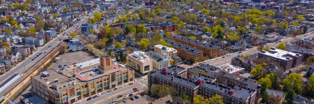 Aerial view of a town near a mental health retreat in Boston, showcasing a mix of urban and suburban features. Buildings, streets, and parking areas are visible amid green foliage. The area includes a railway station with tracks on the left side, offering tranquility and connectivity.