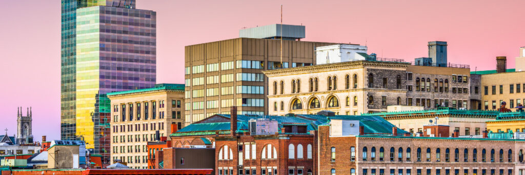 Skyline of a city with a mix of modern and historic buildings under a pink-hued sky, capturing Bostons charm. The architecture includes tall glass and steel structures, evocative of its spirit, alongside older brick buildings—a perfect backdrop for a mental health retreat in the heart of the city.