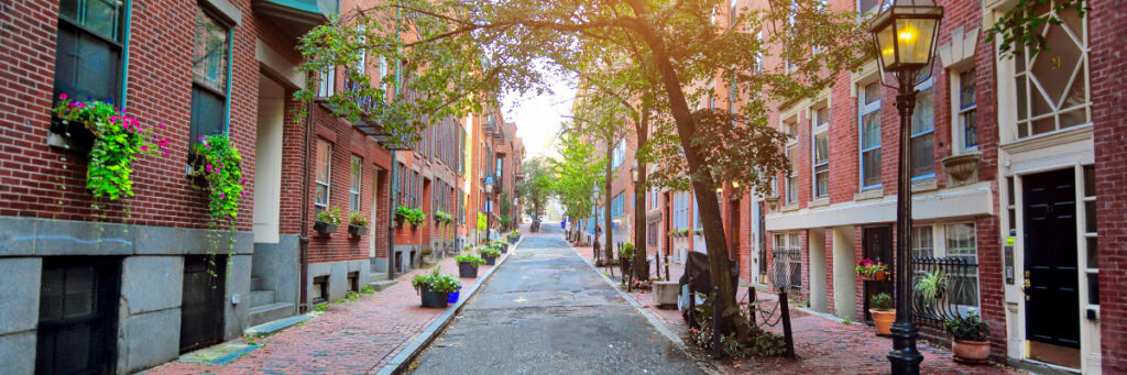 A narrow, cobblestone street in Boston is lined with historic red brick buildings and blossoming flower boxes. Trees arch over the pathway, alongside a vintage lamppost. Sunlight filters through the leaves, creating a warm atmosphere that invites introspection—ideal for a mental health retreat.
