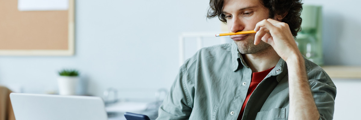 A person with wavy hair is at a desk, contemplating a mental health retreat in Boston on their smartphone. A pencil rests between their lips, and a laptop sits open in front of them. In the background, a corkboard adds to the cozy office or study vibe.