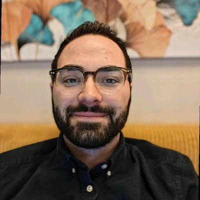 Meet The Staff: A man with dark hair, beard, and glasses is smiling at the camera while sitting on a yellow couch. There is abstract art with blue and brown tones on the wall behind him.