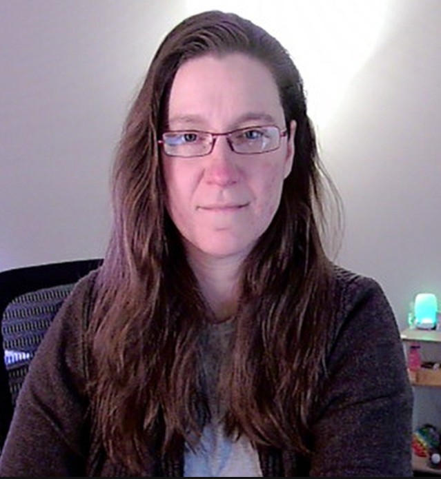 Meet The Staff: A woman with long brown hair and glasses sits indoors, facing the camera and smiling slightly. She is wearing a brown sweater and gray shirt, with softly lit walls and a glowing blue lamp behind her.