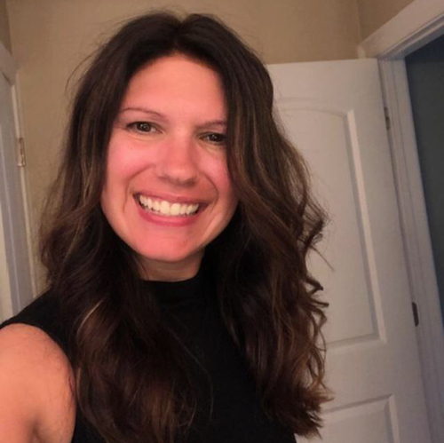 Meet The Staff: A woman with long wavy brown hair and a black sleeveless top smiles at the camera in a well-lit room with an open door in the background.
