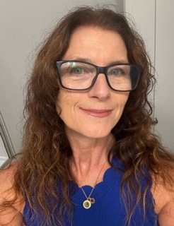 Meet The Staff: A woman with long wavy brown hair, wearing large black-rimmed glasses, a blue sleeveless top, and a pendant necklace, smiles softly while standing indoors against a neutral background.