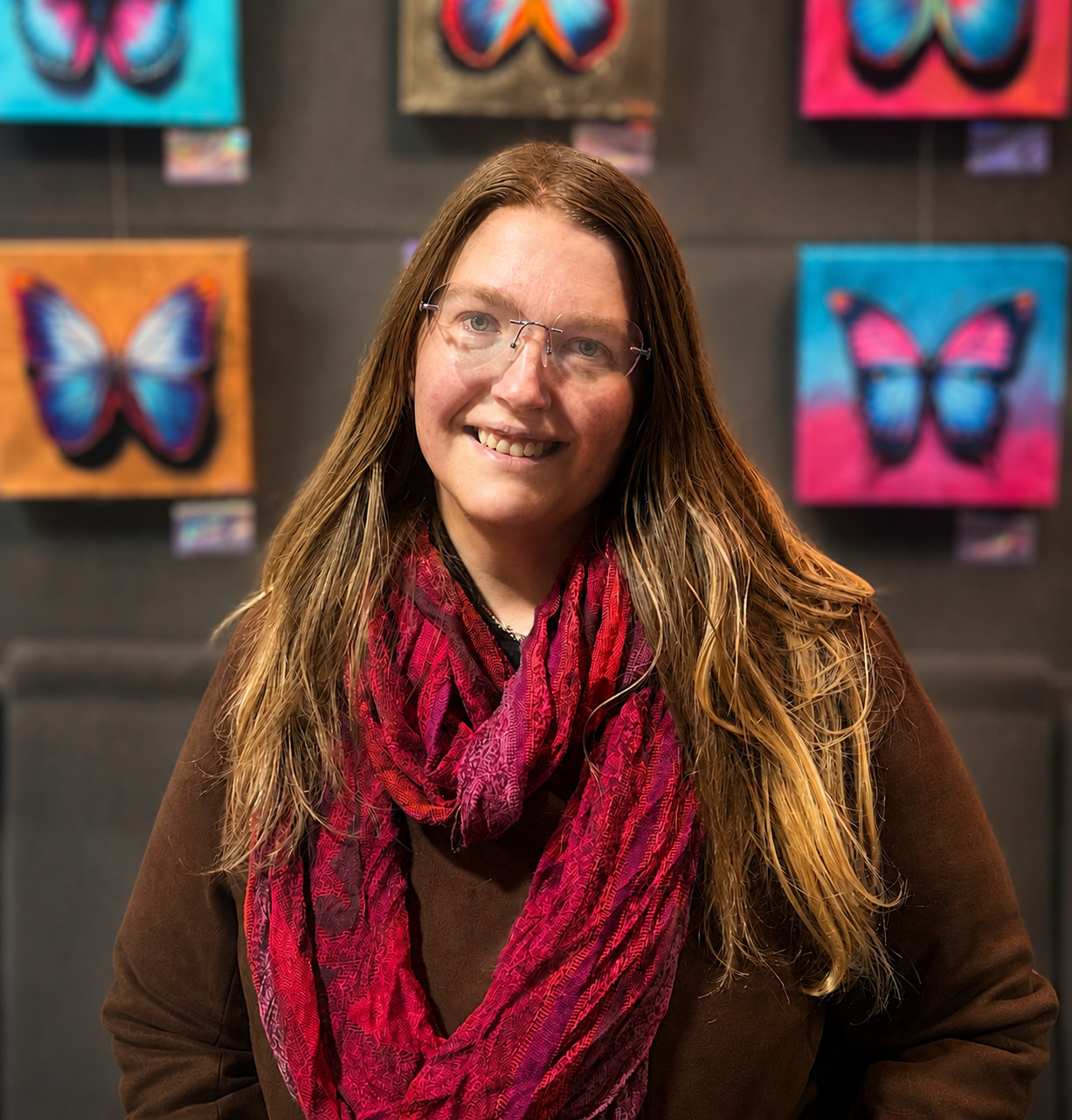 Meet The Staff: A woman with long brown hair and glasses smiles warmly in a red scarf and brown coat, standing before a wall adorned with colorful butterfly artwork.